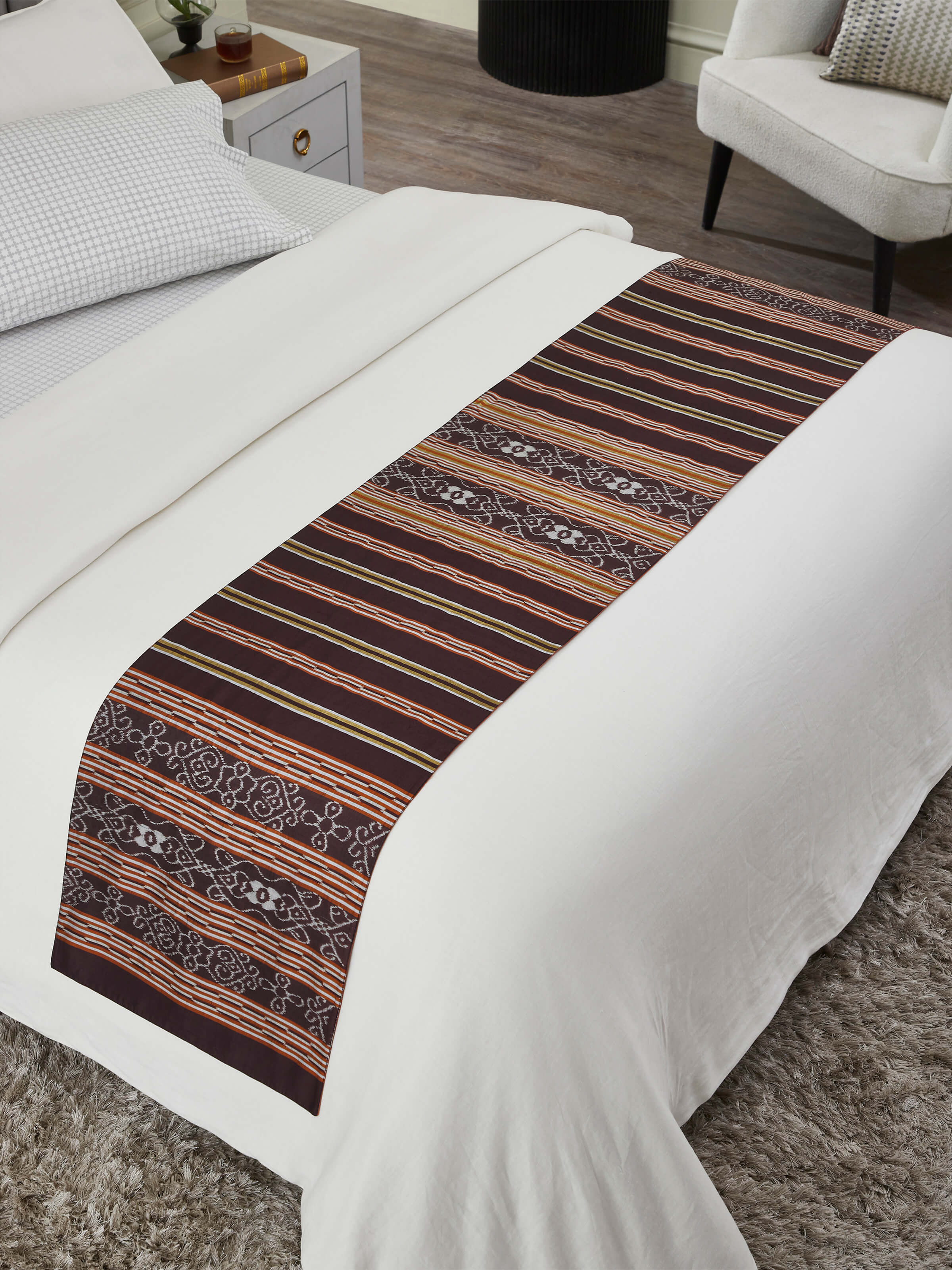 A lifestyle photograph of the Sanchaya Coffee Stripe Bed Runner placed on a bed, showcasing the traditional Odisha handloom craftsmanship and silk Ikat pattern.