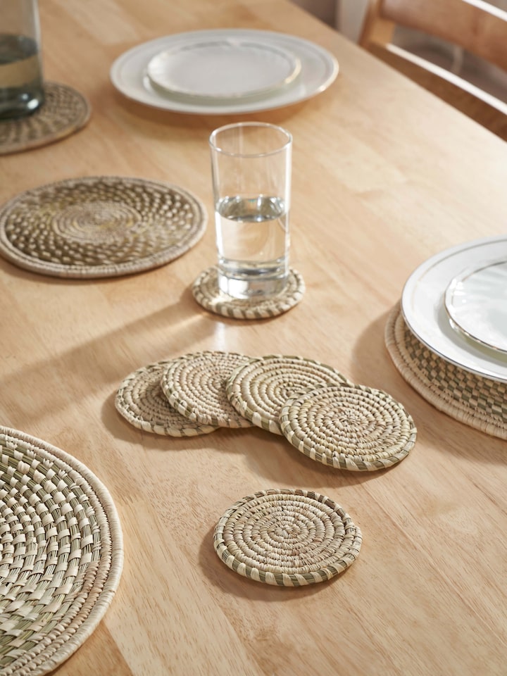 Lifestyle view of Amodini light green Sabai grass coasters arranged under glasses on a rustic wooden table.
